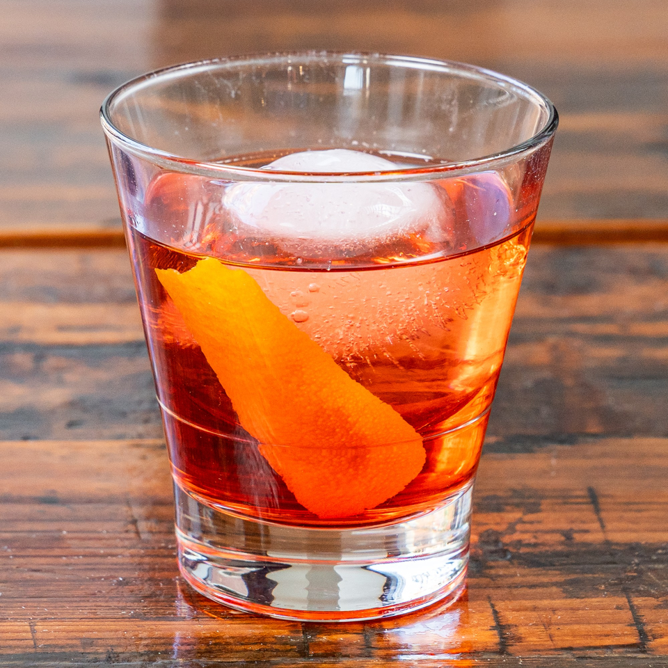 A vibrant orange cocktail with a large ice cube and an orange peel garnish on a wooden table.
