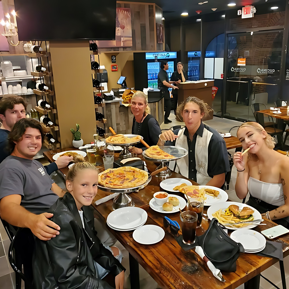 Family and friends enjoying pizzas, burgers, and drinks at a long wooden table in Cucina Pizza & Bar.