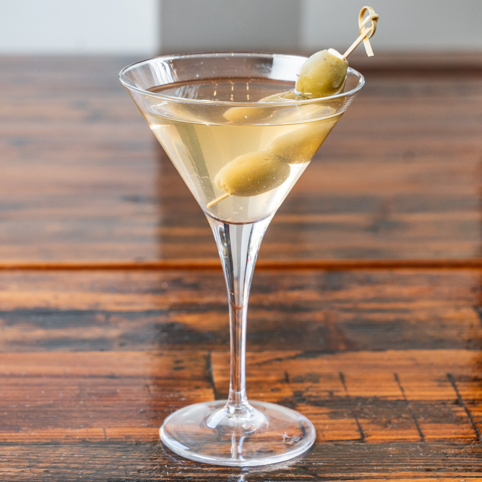 Martini cocktail with three green olives on a skewer, served in a clear glass on a wooden table.