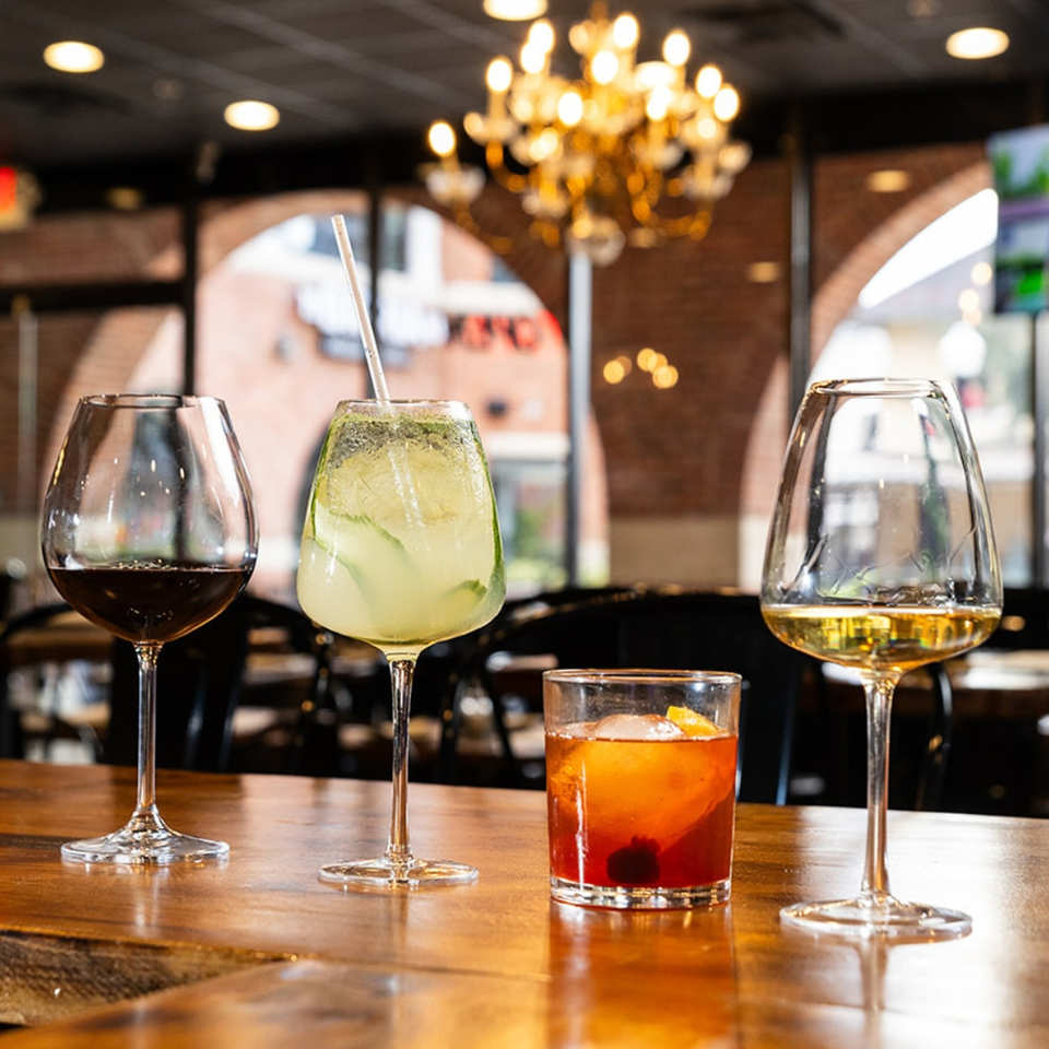 Four drinks, including red wine, a green cocktail, and an Old Fashioned, on a wooden bar.