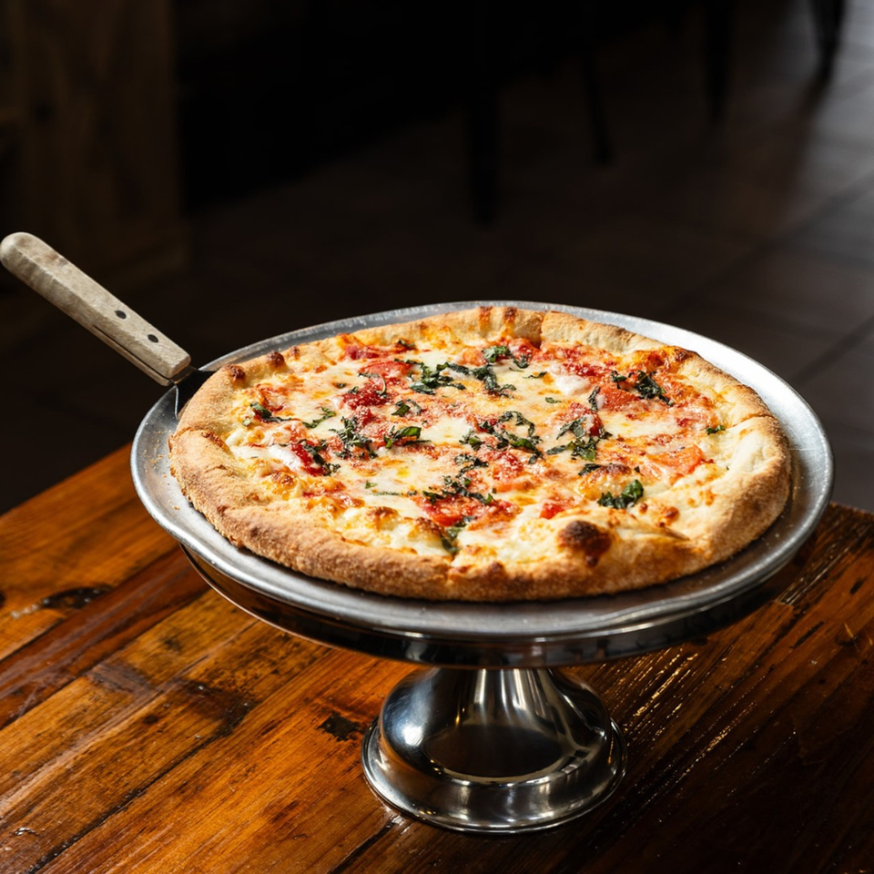A freshly baked Margherita pizza with basil and cheese on a metal stand, on a rustic wooden table.