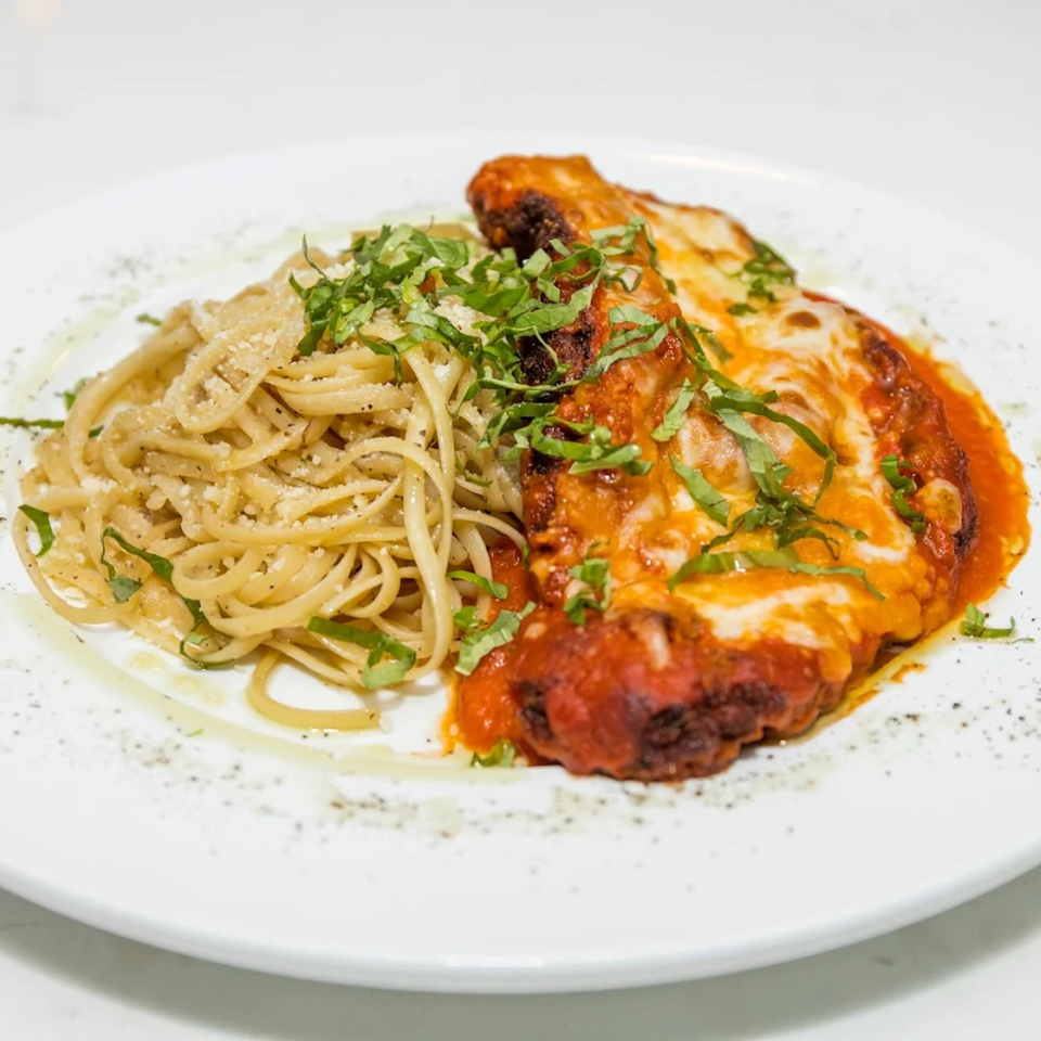 Chicken parmesan with melted cheese and red sauce, served alongside linguine pasta, garnished with fresh basil.