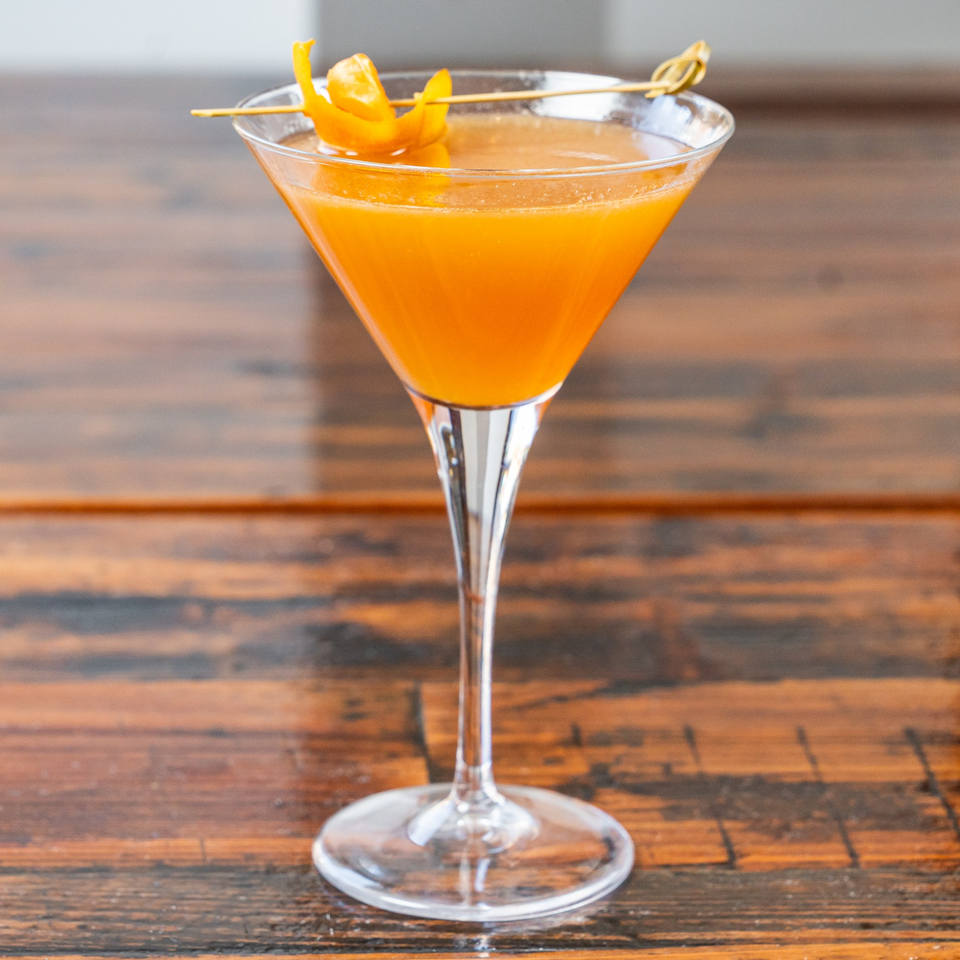 A vibrant orange cocktail in a martini glass, garnished with an orange peel, on a wooden table.