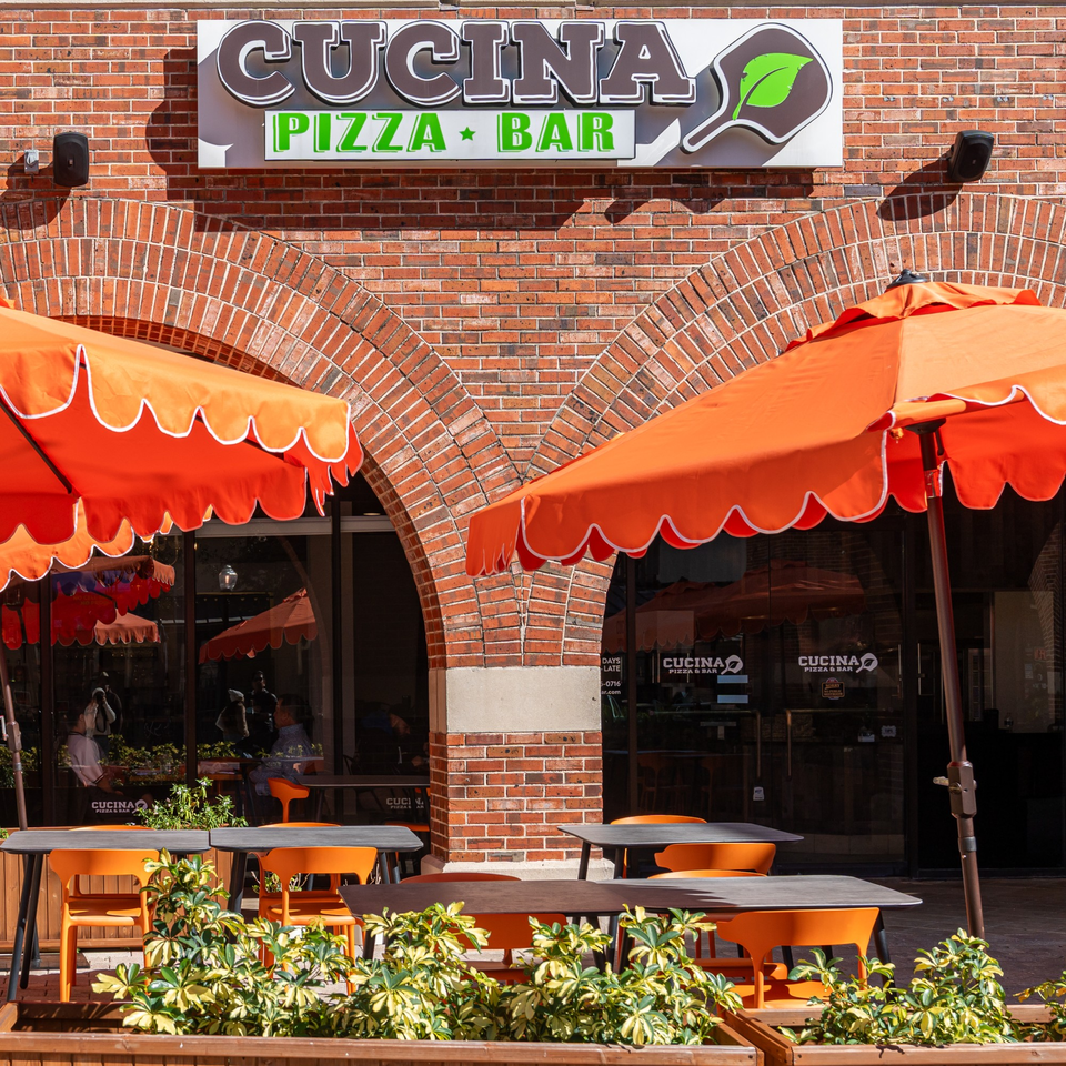 Cucina Pizza & Bar storefront with orange umbrellas, outdoor seating, and a brick facade.
